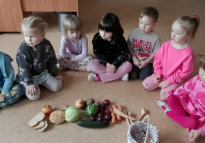 Dzieci siedzą na podłodze przed nimi leżą owoce i warzywa oraz pożywienie, które zjadają kury.