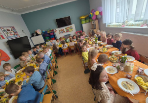 Na zdjęciu dzieci siedzą przy świątecznie udekorowanych stołach podczas wielkanocnego obiadu w przedszkolu. Na stołach znajdują się potrawy i wiosenne dekoracje.