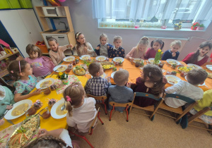 Na zdjęciu dzieci siedzą przy świątecznie udekorowanych stołach podczas wielkanocnego obiadu w przedszkolu. Na stołach znajdują się potrawy i wiosenne dekoracje.