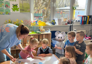 Dzieci siedzą przy stoliku i wraz z nauczycielką i tworzą grupową prace plastyczną