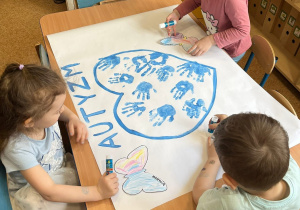 Dzieci siedzą przy stoliku i wraz z nauczycielką i tworzą grupową prace plastyczną