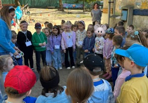 Dzieci z przedszkola stoją w ogrodzie przedszkolnym, w kole i słuchają nauczycielki, która wyjaśnia zasady zabawy