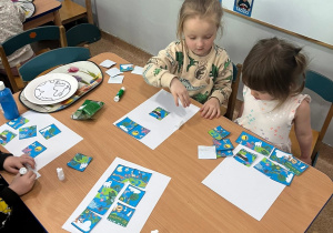 Dzieci siedzą przy stolikach i przyklejają puzzle w temacie Dnia Ziemi na biały karton