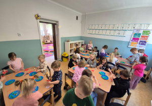 Dzieci siedzą przy stolikach i malują ziemie na talerzach papierowych