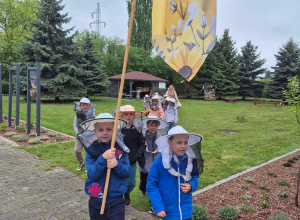 Dzieci idą parami przez teren Pasieki. Na głowach mają kapelusze pszczelarskie. Pierwsza para niesie flagę, na której widnieje pszczoła i plastry miodu