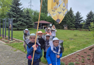 Dzieci idą parami przez teren Pasieki. Na głowach mają kapelusze pszczelarskie. Pierwsza para niesie flagę, na której widnieje pszczoła i plastry miodu