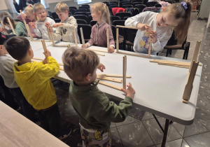 Dzieci stoją przy stole i samodzielnie składają drewniane konstrukcje z przygotowanych elementów podczas warsztatów edukacyjnych.
