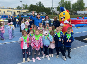 Grupa dzieci z medalami i zielonymi chustami pozuje do wspólnego zdjęcia z Moniką Pyrek na stadionie sportowym wraz z opiekunkami i kolorową maskotką.