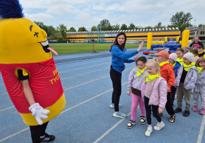 Dzieci ustawione w rzędzie na bieżni stadionu uczestniczą w aktywności sportowej prowadzonej przez instruktorkę, obok kolorowej maskotki wydarzenia.