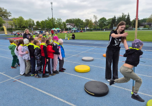 Dzieci ustawione w kolejce uczestniczą w konkurencji sportowej na stadionie, a jedno z nich wykonuje zadanie ruchowe pod opieką instruktorki.