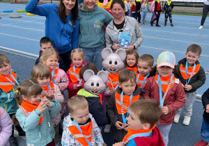 Grupa dzieci z medalami i pomarańczowymi chustami pozuje do wspólnego zdjęcia na stadionie wraz z opiekunkami i kolorową maskotką wydarzenia sportowego.