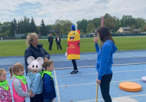 Dzieci stoją na bieżni stadionu i słuchają instrukcji prowadzącej przed rozpoczęciem konkurencji sportowej, a w tle widoczna jest maskotka wydarzenia.