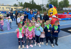 Grupa dzieci z medalami i zielonymi chustami pozuje do wspólnego zdjęcia na stadionie wraz z opiekunkami i kolorową maskotką podczas wydarzenia sportowego.