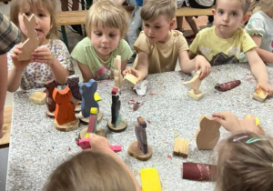 Dzieci siedzą przy podłużnym stole a na środku stołu wystawione są kolorowe drewniane zabawki