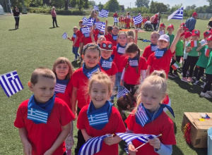 Grupa dzieci w czerwonych koszulkach i niebieskich apaszkach z flagą Grecji pozuje do zdjęcia na stadionie ChKsu