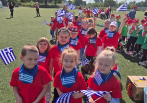 Grupa dzieci w czerwonych koszulkach i niebieskich apaszkach z flagą Grecji pozuje do zdjęcia na stadionie ChKsu