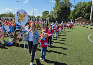 Dzieci idą w parach za nauczycielem, nauczyciel niesie stand z logo przedszkola. Zdjęcie jest robione na stadionie ChKs'u.