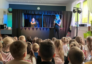 Zdjęcie przedstawia dzieci siedzące tyłem na dywanie oraz scenę Teatru Pacuś.