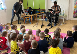 Zdjęcie przedstawia dzieci siedzące tyłem na dywanie oraz dwóch muzyków grających na instrumentach
