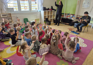 Zdjęcie przedstawia dzieci siedzące na dywanie i klaszczące z rękami do góry. Na scenie widać dwóch muzyków, jednego grającego na gitarze i drugiego klaszczącego.