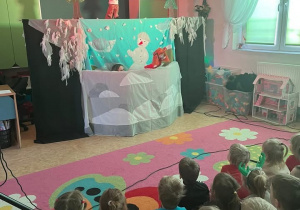 Zdjęcie przedstawia dzieci siedzące tyłem na dywanie oraz scenę Teatru Pacuś.