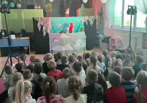 Zdjęcie przedstawia dzieci siedzące tyłem na dywanie oraz scenę Teatru Pacuś.