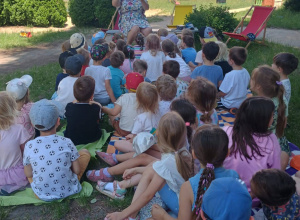 Zdjęcie przedstawia dzieci siedzące tyłem na kocach w ogrodzie przedszkola i słuchają bajki czytanej przez nauczyciela.