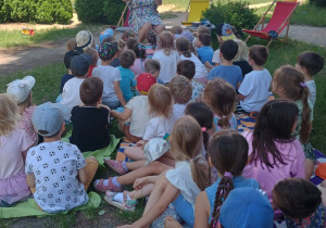 Zdjęcie przedstawia dzieci siedzące tyłem na kocach w ogrodzie przedszkola i słuchają bajki czytanej przez nauczyciela.