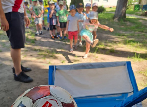 Dzieci biorą udział w sportowej zabawie na świeżym powietrzu, wykonując strzały piłką do małej bramki pod okiem prowadzącego. Pozostałe przedszkolaki z zainteresowaniem czekają na swoją kole