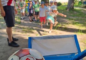 Dzieci biorą udział w sportowej zabawie na świeżym powietrzu, wykonując strzały piłką do małej bramki pod okiem prowadzącego. Pozostałe przedszkolaki z zainteresowaniem czekają na swoją kole