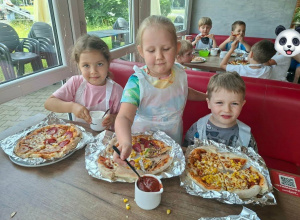 Trójka dzieci siedzi przy stoliku a przed nimi na talerzu leży pizza wykonana przez nich.