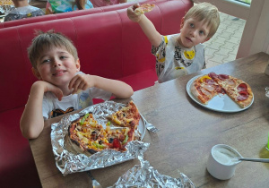 Dzieci siedzą przy stołach w pizzerii i próbują pizz wykonanych przez siebie.