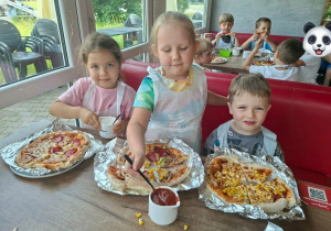 Dzieci siedzą przy stołach w pizzerii i próbują pizz wykonanych przez siebie.