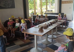 Dzieci siedzą przy stołach w pizzerii, na środku jest stół z przygotowanymi produktami. Pan w żółtej bluzce tłumaczy dzieciom proces robienia pizzy.