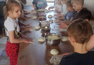 Dzieci stoją przy długim stole z przygotowanymi produktami i przygotowują się do robienia pizzy.