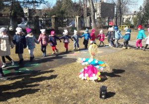 Dzieci w ogrodzie przedszkolnym tańczą w dużym kole i śpiewają wiosenną piosenkę. W środku koła stoi Marzanna.
