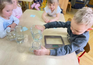 Dzieci wykonują eksperyment z wodą. Ich zadaniem jest sprawdzenie w którym słoiku jest więcej wody. Słoiki są różnych kształtów. Dzieci przelewają wodę do szklanki i okazuje się ze wszędzie jest tyle samo wody.