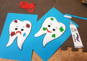 Dwie ilustracje zębów leżą na podłodze. Na jednej ilustracji ząb jest dziurawy i smutny, na drugiej ząb jest wesoły i załatany plombami z plasteliny. Obok leżą papierowe sylwety pasty do zębów i szczoteczki.