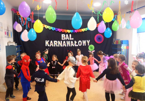 Dzieci z grupy średniaków w strojach karnawałowych trzymając się za ręce tańczą w kole w swojej sali. Sala przystrojona balonami i kolorowymi dekoracjami. W tle napis BAL KARNAWAŁOWY
