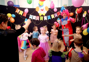 Dzieci z najmłodszej grupy i Nauczycielki w strojach karnawałowych tańczą w sali.Dookoła kolorowe dekoracje, balony i serpenytyny.