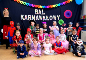 Zdjęcie grupowe dzieci z nauczycielką z grupy 4-5 - latków. Wszyscy ubrani w stroje karnawałowe. W tle na ciemnym tle napis BAL KARNAWAŁOWY wokół kolorowe papierowe ozdoby..