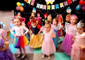 Zdjęcie grupowe. Dzieci z najmłodsszej grupy tańczą na balu. Wszystkie stoją i trzymają rączki na biodrach. Dzieci w strojach karnawałowych. W tle dekoracja - klaun z papieru, napis BAL KARNAWAŁOWY, kolorowe balony i serpentyny.