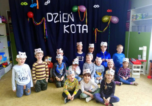 Zdjęcie grupowe. Dzieci z grupy najstarszej pozują w własnoręcznie zrobionych opaskach z obrazkiem na głowach na tle dekoracji z napisem DZIEŃ KOTA.