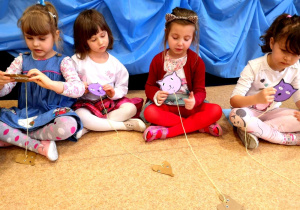 Dziewczynki z grupy 4-5 - latków siedzą na podłodze w kole, biorą udział w konkursie polegającym na tym kto szybciej nawinie nitkę na sylwetę w kształcie kota. Na końcu nitki znajduje się papierowa sylweta w kształcie myszy.