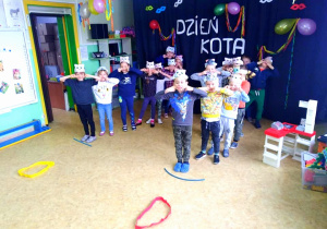 Dzieci z najstarszej grupy biorą udział w konkurencji sportowej. Stoją ustawieni w dwa rzędy, przed nimi na podłodze ułożone są kolorowe szarfy.