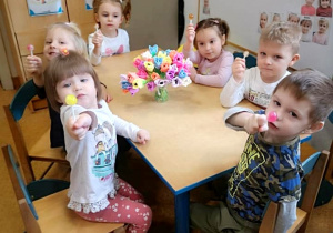 Dzieci z grupy najmłodszej siedzą przy stoliku. Jedzą lizaki. Na stole stoi bukiet kolorowych kwiatów w wazonie.