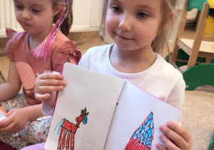 Dziewczynka siedzi skrzyżnie na podłodze, prezentuje lasnoręcznie narysowane obrazki w zrobionej przez siebie książce. Na jednej stronie narysowany konik, na drugiej domek.