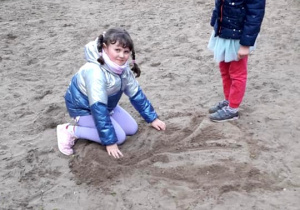 Dziewczynki w ogrodie przedszkolnym bawią się ziemią. Jedna rysuje na ziemi drzewo, druga ogląda obrazek.