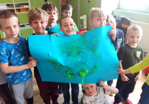 Grupa chłopców prezentuje swoją pracę plastyczną. Na niebieskim papierze narysowana kula ziemska ozdobiona papierem kolorowym.