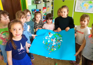 Grupa dziewczynek prezentuje swoją pracę plastyczną. Na niebieskim papierze narysowana kula ziemska ozdobiona papierem kolorowym i gazetami.